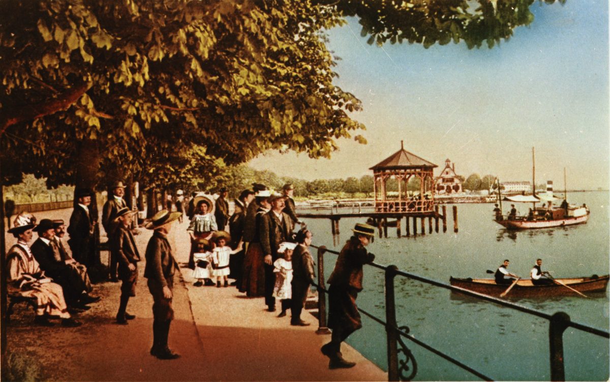Kolorierte Postkarte um 1900: Spaziergänger unter Bäumen auf der Bregenzer Seepromenade, dahinter der Fischersteg mit seinem Pavillon und Ruderboote auf dem See